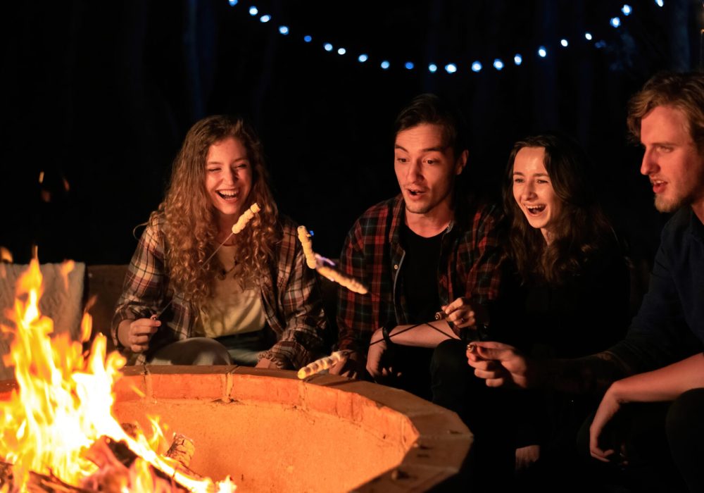 A group of happy young friends near a campfire at glamping, night. Two men and women. Frying marshmallow